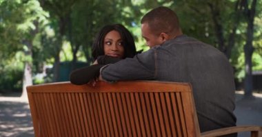 Sevimli çift halka açık bir parkta bir bankta otururken birbirlerine sevgiyle bakar. Mutlu Afrikalı erkek ve dişi, günü parkta geçirirken romantik bir şekilde göz göze gelirler. 4k