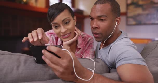 一个黑人和一个女人一边听音乐一边用他们的智能手机 一对非洲裔美国夫妇坐在沙发上使用他们的手机 — 图库视频影像