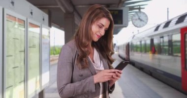 Tren istasyonundayken telefonundan e-posta okuyan beyaz bir kadın. Güzel, modern bir beyaz kadın bir tren istasyonunda cep telefonunu kontrol ediyor. 4k
