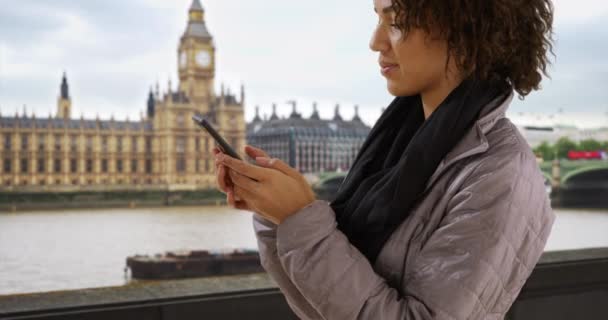 ビッグ クロック タワー近くのモバイル デバイスに関する20代のテキストのかわいい黒人女性 ロンドンのスカイラインを眺めながら携帯電話で友達にヨーロッパを旅行する100万人の女の子 — ストック動画