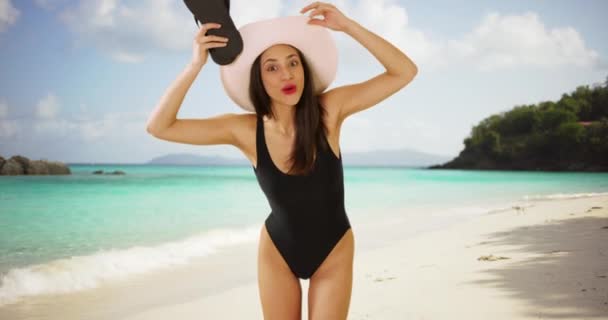 一个拉丁女孩在海滩上摆出一副滑稽的姿势要拍一张肖像 一个西班牙女人在海滩上拿着帽子和鞋子玩得很开心 — 图库视频影像
