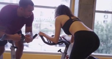 Spor salonundaki çift durağan bisiklete binip pedal çeviriyor. Siyah adam ve İspanyol kadın.