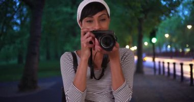 Dijital kameralı siyahi kadın halka açık bir parkın yakınında çekim yapıyor. Siyah kadın fotoğrafçı az önce kamerasıyla çektiği bir fotoğrafı kontrol ediyor. 4k