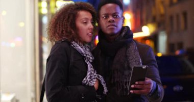 Kayıp Afro-Amerikalı çift, akıllı telefondan şehir caddesinde yol tarifi için harita kullanıyor. Milenyum siyahi erkek ve kadın geceleri şehirde cep telefonu uygulaması kullanıyor. 4k
