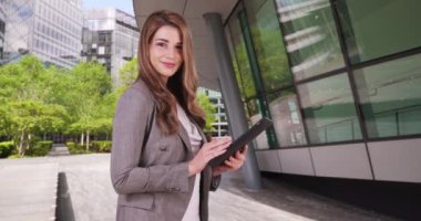 Ofis binasının dışında tablet üzerinde çalışan güzel beyaz profesyonel bir kadın. Beyaz kadın bir ped kullanıyor ve kameraya gülümsüyor. 4k