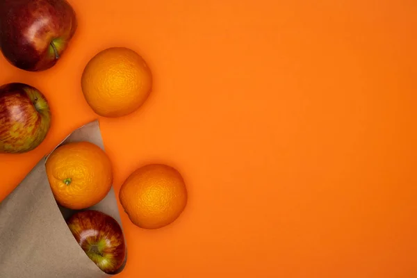 Свіжі червоні яблука та апельсини в паперовій упаковці на яскраво-помаранчевому фоні — стокове фото