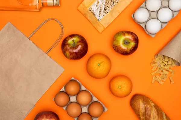 Паперовий еко-пакет, пляшка оливкової олії, яйця, хліб, боби, червоні яблука, фрукти, спіральна паста на помаранчевому фоні — стокове фото