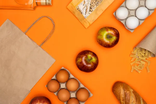 Паперовий еко-пакет, пляшка оливкової олії, яйця, хліб, боби, червоне яблуко, спіральна паста на помаранчевому фоні — стокове фото
