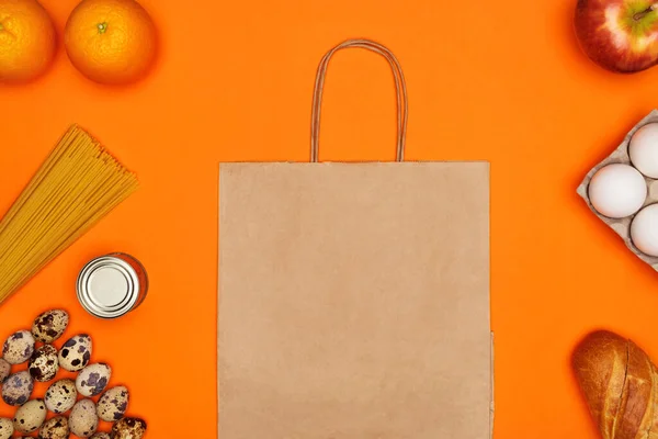 Паперова еко сумка, яйця, спагетті, хліб, червоне яблуко, апельсини та металева олова на яскраво-оранжевому ізольованому фоні — стокове фото