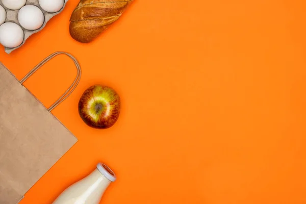 Паперова екологічна сумка, скляна пляшка молока, хліб, білі яйця, червоне яблуко на помаранчевому ізольованому фоні — стокове фото