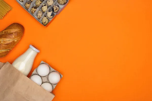 Білі яйця і пляшка молока в паперовій еко сумці. Спагетті, хліб і перепелині яйця на помаранчевому фоні — стокове фото