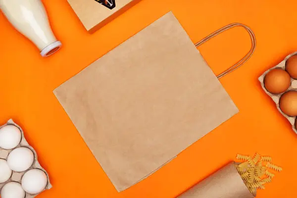 Паперовий еко сумка, скляна пляшка молока, яйця, спіральна паста та коробка чаю на яскраво-оранжевому ізольованому фоні — стокове фото
