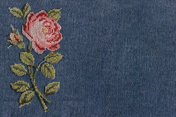 abstract background cross-stitched pink rose on the background texture blue denim fabric closeup