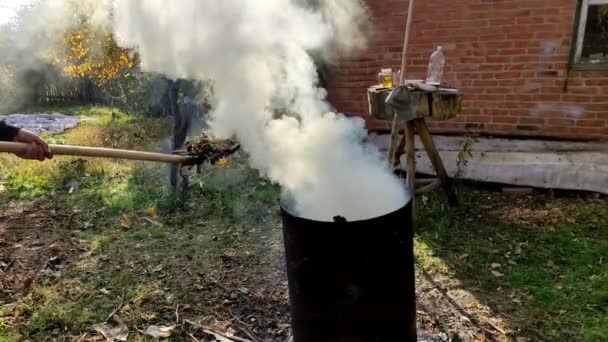 燃えるような落ち葉の煙とバレル 男はピッチフォークでそれに葉をスロー 4Kビデオ — ストック動画