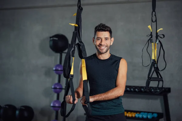 Young man stands in a gym holding suspension equipment looking at camera.