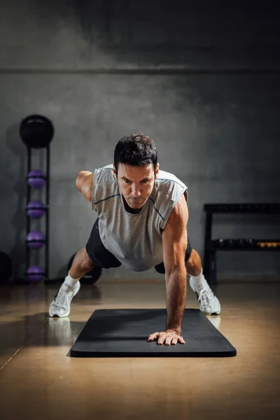 Strong caucasian man looks down while does push-ups with one hand in a dark gym.