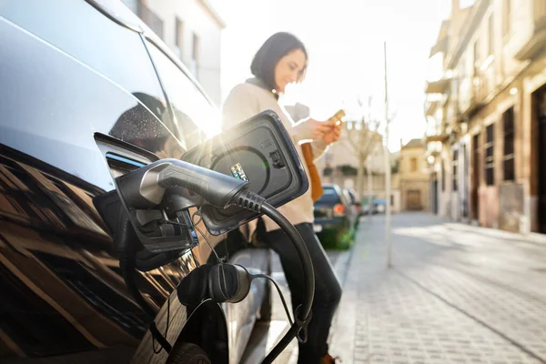 Vrouw die wacht op elektrische auto om op te laden op de parkeerplaats bij zonsondergang. — Stockfoto