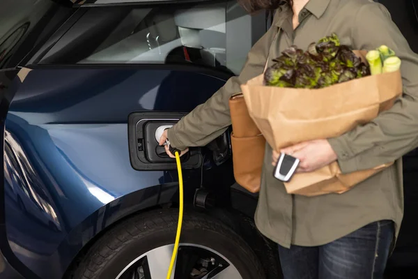 Vrouw pluggen een lader om elektrische auto stopcontact thuis na het winkelen boodschappen. — Stockfoto