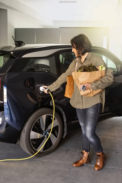食料品を持つ女性は自宅で電気自動車用ソケットへの充電器を差し込みます. — ストック写真