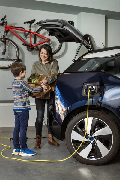 Vrouw en jongen halen de boodschappen terwijl elektrische auto is opladen thuis. — Stockfoto