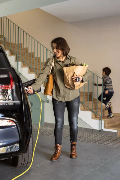 Vrouw met boodschappen stekkers een lader om elektrische auto stopcontact thuis. — Stockfoto