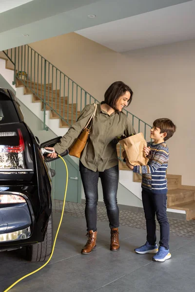 Lachende moeder en zoon opladen elektrische auto thuis. — Stockfoto