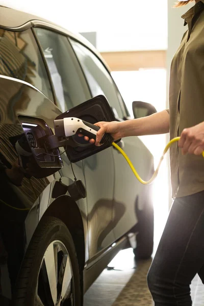 Vrouw plugs een oplader naar elektrische zwarte auto socket thuis garage. — Stockfoto