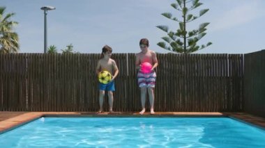 Güneşli bir günde, bir topla oynayıp mavi bir su havuzuna atlayan iki çocuk. Kardeşler yarışıyor. 4k video, yavaş çekim.