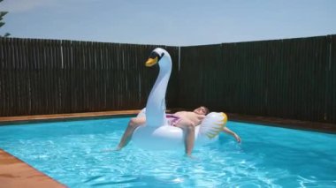 Genç mutlu çocuk mavi berrak havuzda şişme kuğu oyuncağında rahatlıyor. Bir yaz günü, havuzda kuğu şamandırasıyla güneşlenen bir çocuk. 4k video, yavaş çekim.