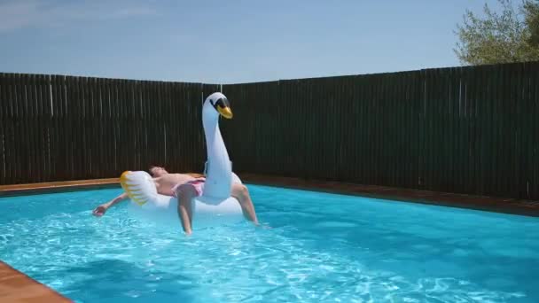 ティーン男の子リラックスオンインフレータブル白鳥のおもちゃでブルークリアプール 休暇中の夏の日に白鳥の群れと一緒にスイミングプールで日光浴 4Kビデオ スローモーション — ストック動画