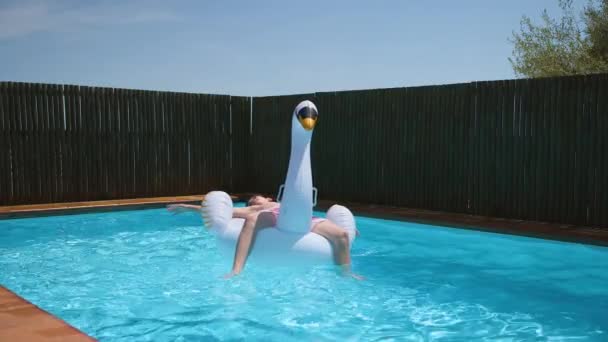 ティーン男の子リラックスオンインフレータブル白鳥のおもちゃでブルークリアプール 子供は休暇中の夏の日に白鳥の群れと一緒にスイミングプールで遊びます 4Kビデオ スローモーション — ストック動画