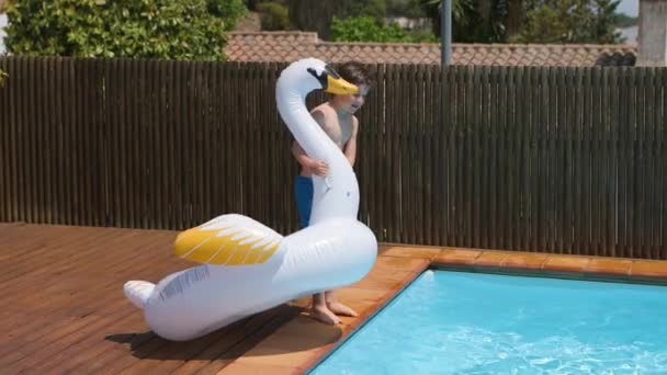 少年はジャンプし 青いプールで膨脹可能な白鳥のおもちゃで遊ぶ 子供は休暇中の夏の日にスイミングプールの白鳥の浮き輪から滑ったり落ちたりします 4Kビデオ スローモーション — ストック動画