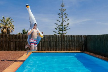 Komik komik çocuk kafasında şişme bir kuğuyla havuza atlıyor. Çocuk özel bir evin arka bahçesinde havuzda eğleniyor. Tatil, yaz tatili. Kopyalama alanı