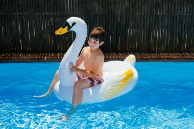 Yaz tatili kavramı. Çocuk kristal mavi suyla dolu bir havuzda şişme bir kuğu yüzüğünde oynuyor. Çocuk bir hayvan yatağında güneşli bir günün tadını çıkarıyor. Yatay