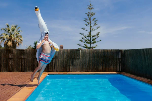 Funny hilarious boy jumps into a pool with an inflatable swan on his head. Kid having fun in a pool of a private house backyard. Vacation, summertime. Horizontal copy-space