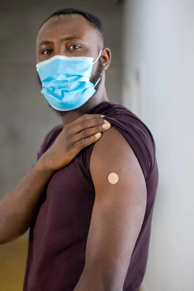 African American man with protective mask looking at camera proudly showing adhesive bandage plaster on arm after getting the covid 19 vaccination standing in a clinic during coronavirus outbreak