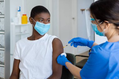 Genç Afrikalı Amerikalı kadın doktor muayenehanesinde oturuyor ve koluna grip aşısı oluyor. Koruyucu üniformalı hemşire, lateks eldivenler ve yüz aşısı, klinikteki Afrikalı Amerikalı kadın hastayı ürküttü.