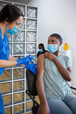 Koruyucu üniformalı, Afrika kökenli Amerikalı genç bir kadına aşı uygulayan, bir hastanenin bekleme odasında oturan bir sağlık çalışanı. Sağlık sigortası aşısı ve sağlık hizmetlerini hesaba katın. Dikey
