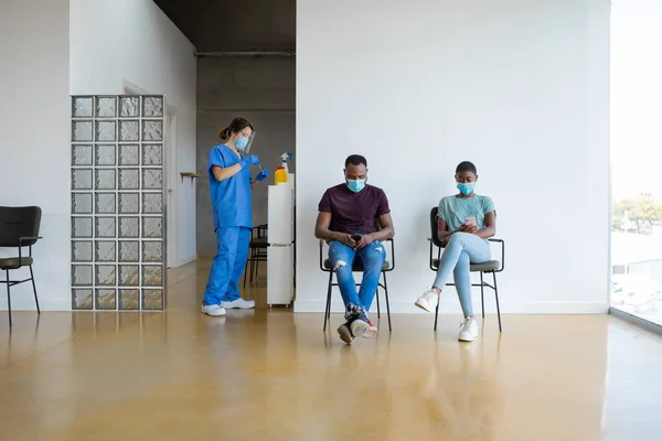 Young African American female and male patients browsing on smartphone, sitting in a waiting room near female doctor in protective uniform, latex gloves and face mask, before vaccinating in clinic.