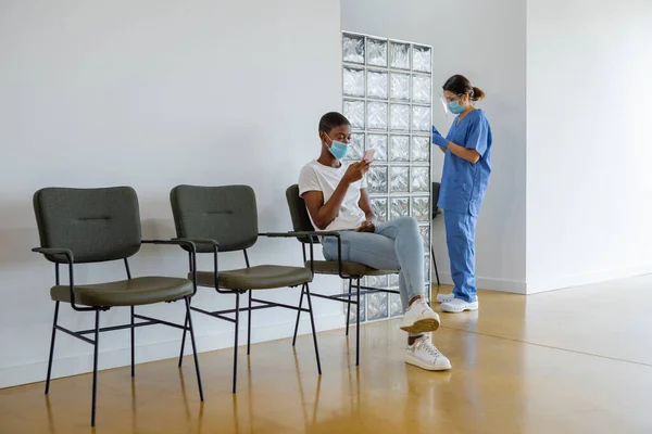 Young African American female patient using smartphone while sitting in a waiting room. Female health worker in protective uniform, latex gloves and face mask standing in clinic.