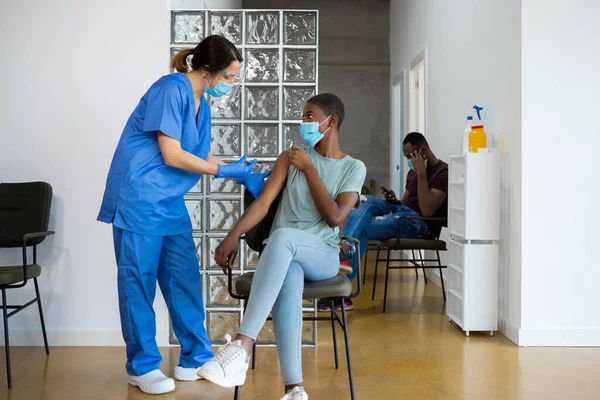Health female worker with protective uniform applying vaccine to a young African American woman, sitting in a waiting room of a hospital. Concept healthcare vaccination and medical services.