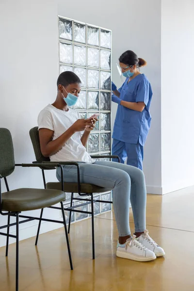 Young African American female patient using smartphone while sitting in a waiting room. Female health worker in protective uniform, latex gloves and face mask standing in clinic.