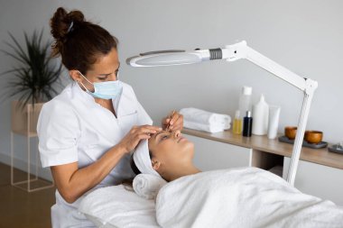 Güzellik uzmanından cımbızla kaplıca salonunda büyüteç altında kaş kılı çıkaran bir kadın.