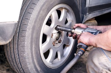 Benzin istasyonundaki bir tamirci, araba tekerleğinin yerine bir işçinin elleri siyah yakıt yağında ve direksiyona yakın vidaları sökmek için bir araç kullanıyor. Lastik servisi, iş akışı