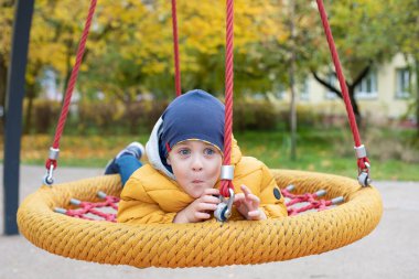Parlak ceketli mutlu bir çocuk salıncağa biniyor. Tatmin oldu. Salıncak sepeti, hamak. Çocukluk kavramı.