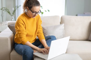 Evdeki kanepede dizüstü bilgisayar kullanan bir kadın. Rahat bir çalışma ortamı, serbest çalışan.