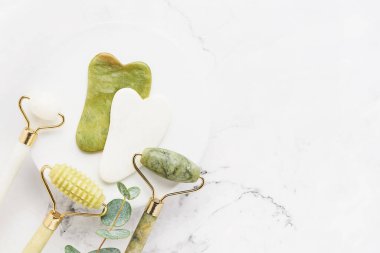 Masaj aletleri. Gua Sha masaj aleti, yeşim yüz silindirleri ve mermer arka planda okaliptüs dalı.