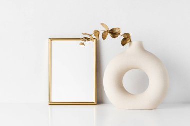 Altın çerçeve maketi ve yuvarlak seramik vazo. Beyaz bir masanın üzerinde altın renkli okaliptüs dalı var.
