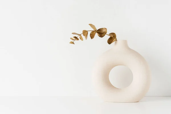 Kulatá váza se zlatou eukalyptovou větví na bílém stole u zdi. Stock Obrázky