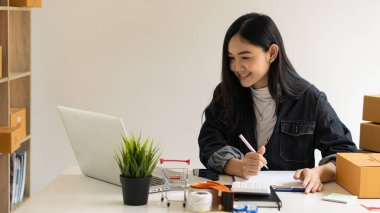 Güzel Asyalı KOBİ işletme sahibi online alışverişte dizüstü bilgisayarda çalışıyor ve evdeki masada paket kutuları var - KME çevrimiçi iş fikri ve kutu teslimatı.